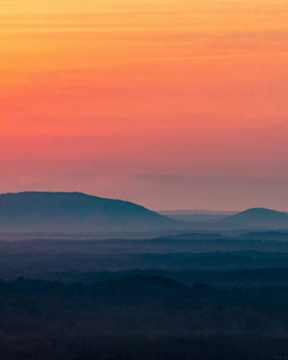 Sunset over layered mountain ridges, warm orange and pink sky above hills fading into mist